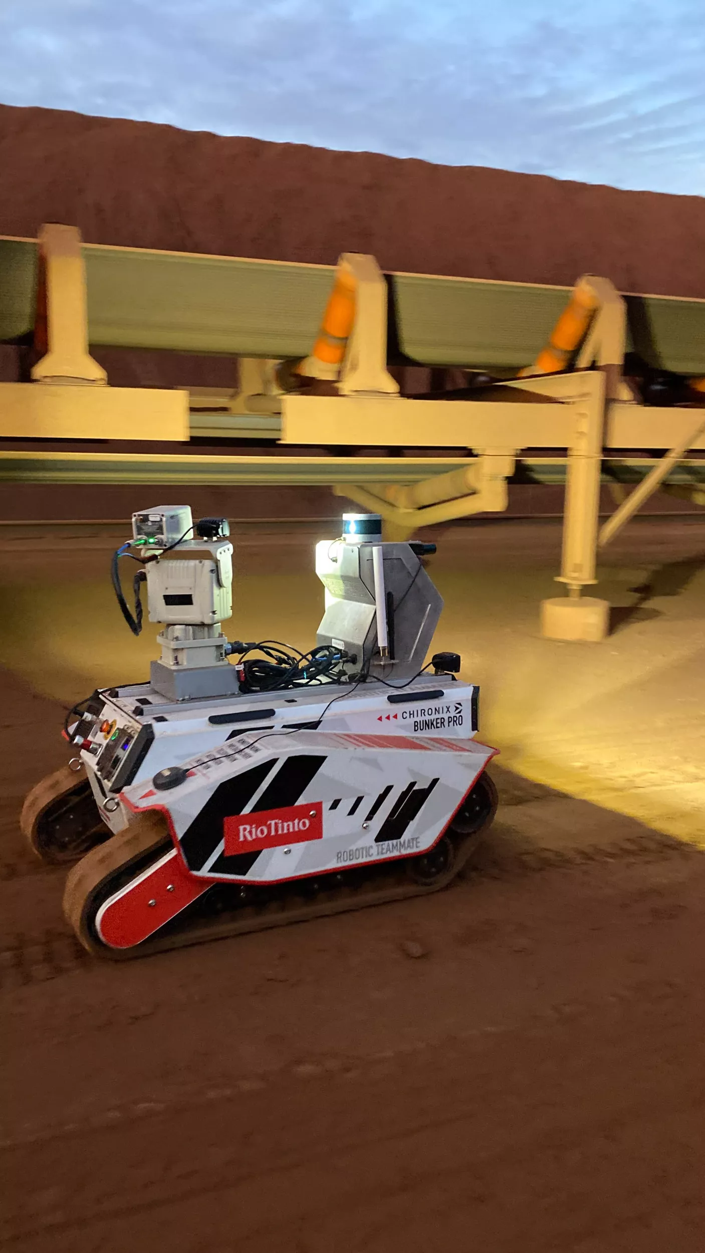 Tracked inspection robot beneath a mine conveyor at dusk